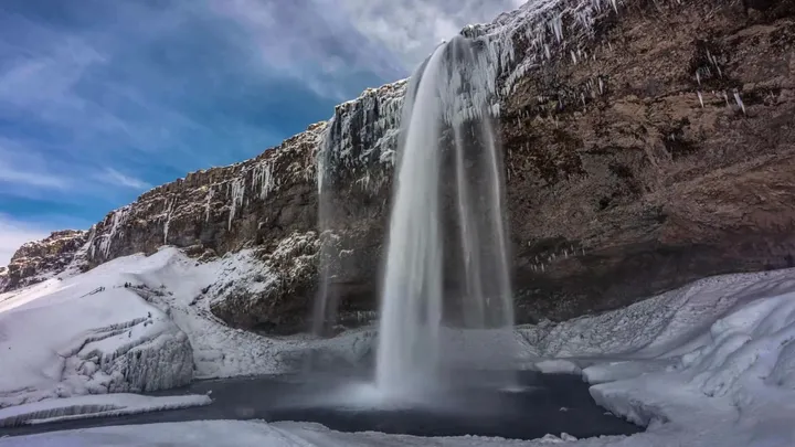 A still photo of an Icelandic waterfall in winter — frozen mid-pour against snow and ice